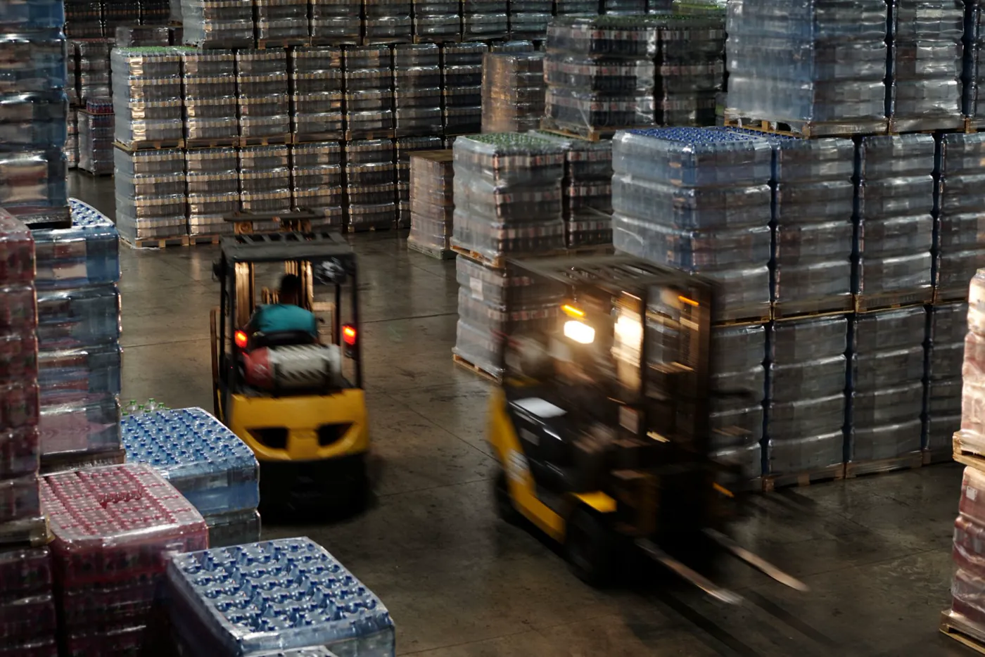 Empilhadeiras em movimento entre pilhas de paletes de bebidas em armazém de centro de distribuição