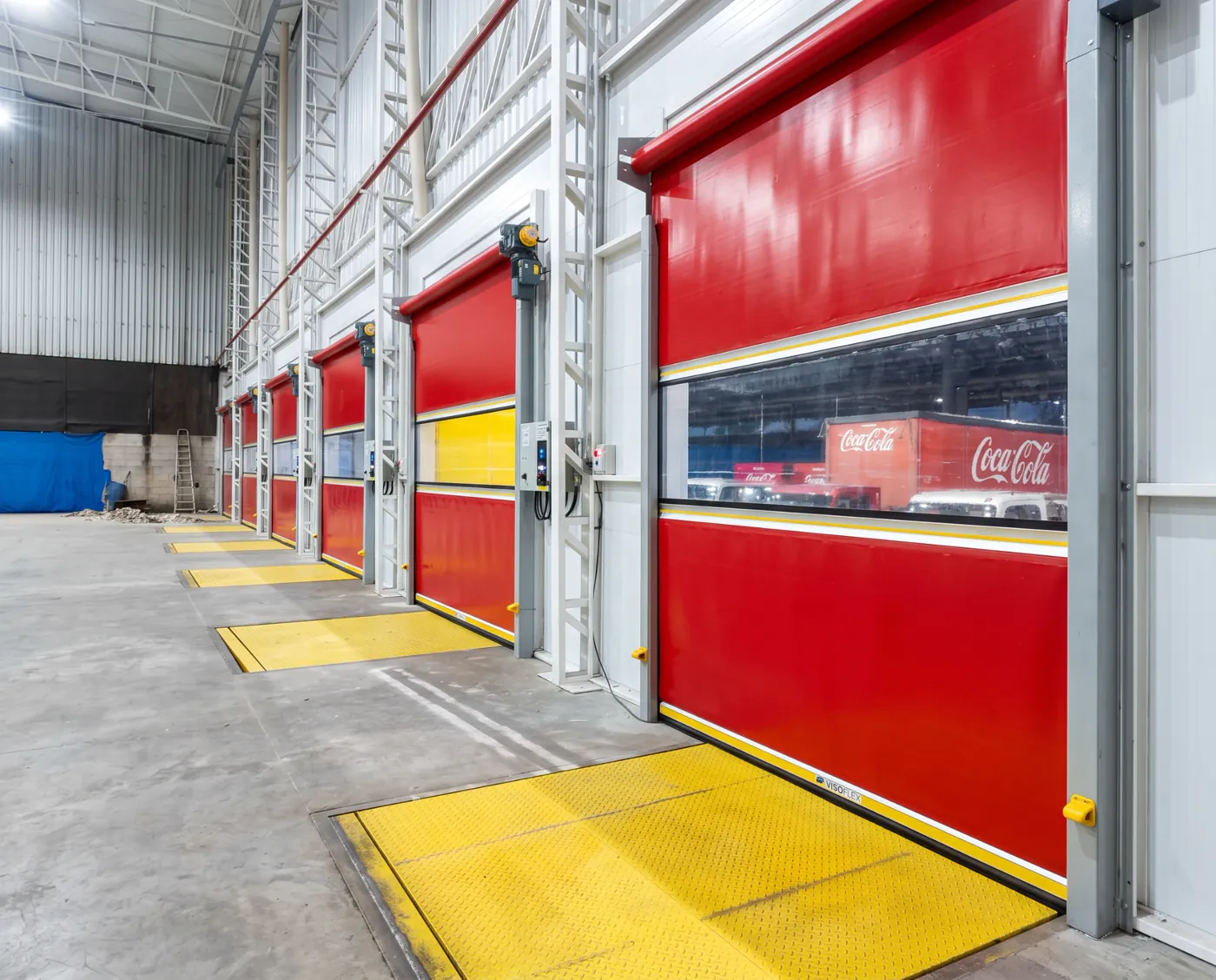 Sequência de portas rápidas Visoflex VFX103 Dock vermelhas com visor nas docas de expedição da Coca-Cola, com niveladores amarelos e caminhões da marca do lado de fora