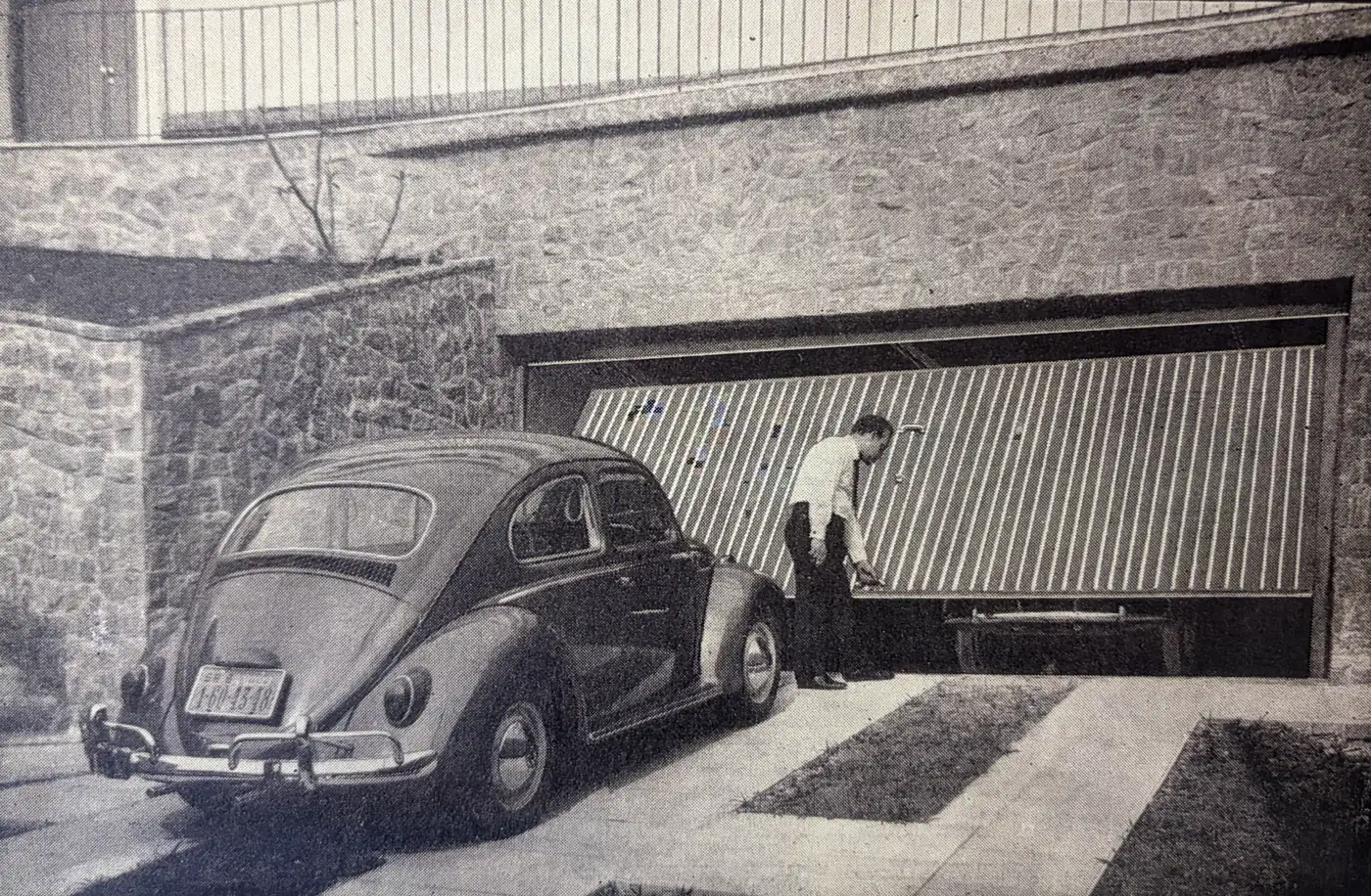 Foto histórica em preto e branco de um homem abrindo uma porta basculante de garagem, com um Fusca estacionado em frente