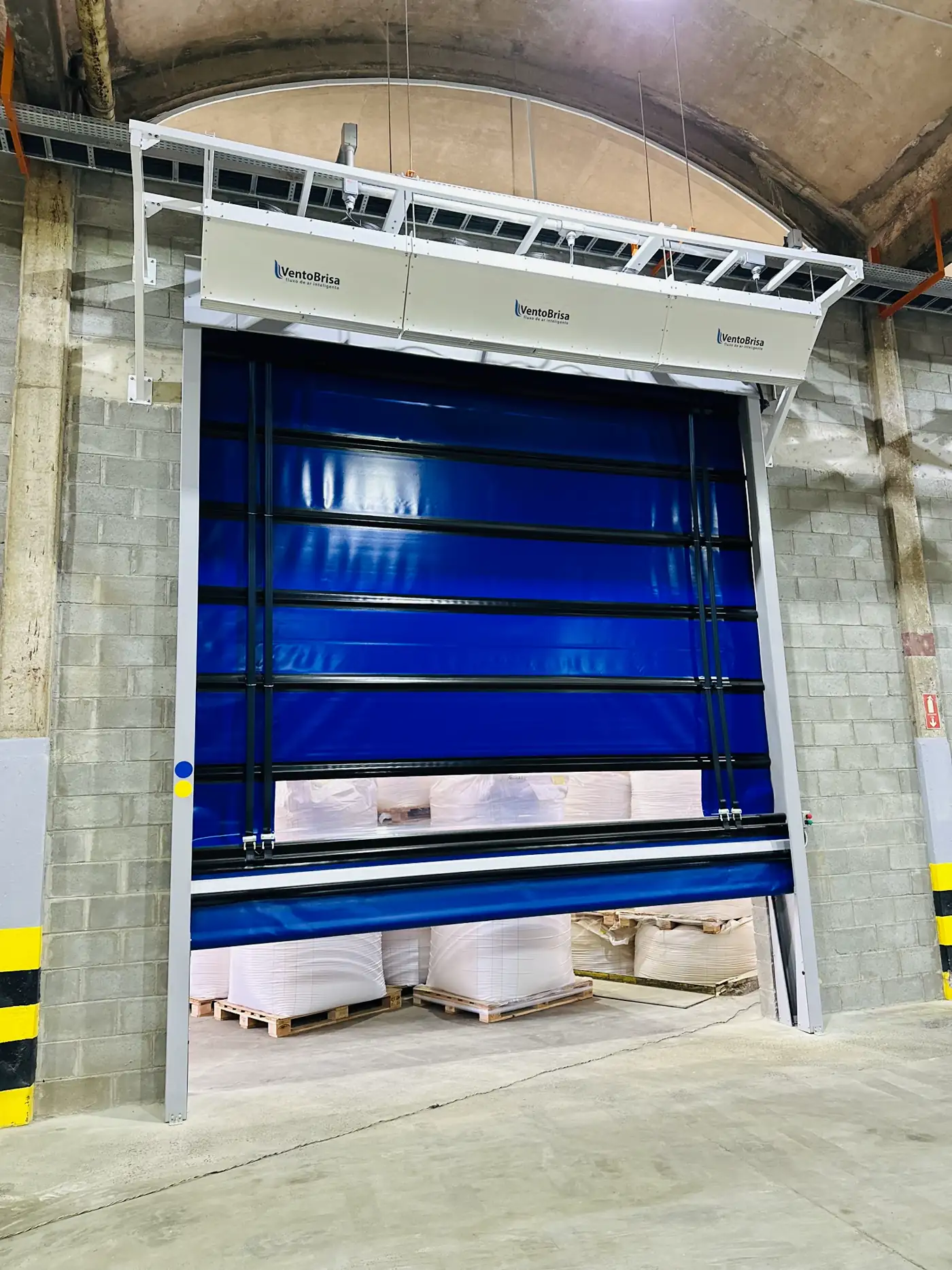 Cortina de ar instalada sobre porta rápida azul aberta, com paletes ao fundo