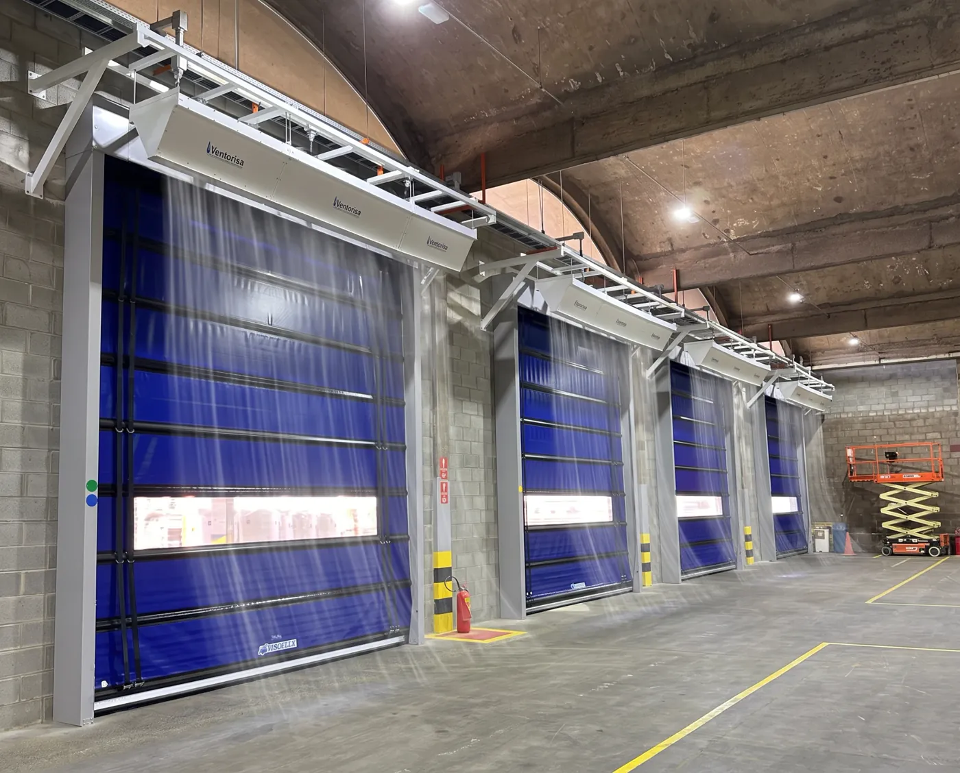 Sequência de cortinas de ar industriais sobre portas rápidas azuis em galpão, com o fluxo de ar descendente ilustrado