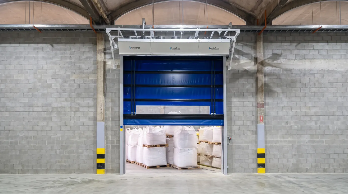 Porta rápida Visoflex VFX501 azul com cintas e visor abrindo grande vão de armazém de big bags, com cortina de ar instalada acima